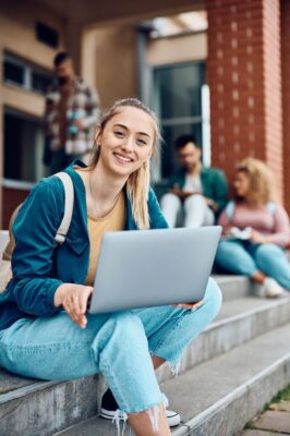 Junge Studentin mit Laptop sitzt lächelnd auf den Stufen vor einem Universitätsgebäude